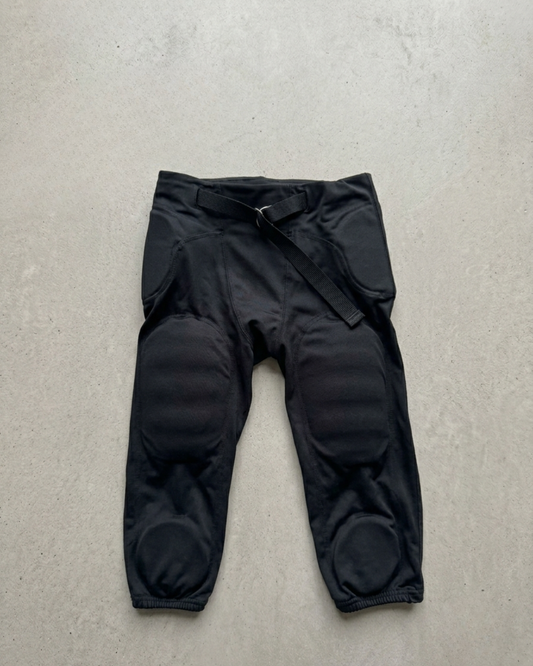 Black padded football pants laid flat on a pale background, integrated thigh and knee pads, webbing belt at the waist.
