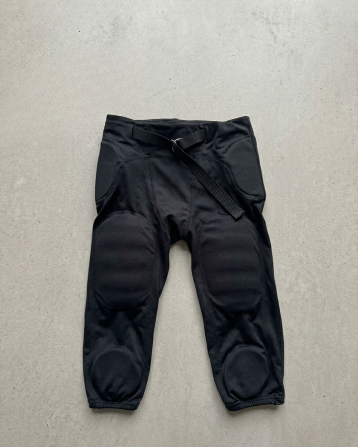 Black padded football pants laid flat on a pale background, integrated thigh and knee pads, webbing belt at the waist.